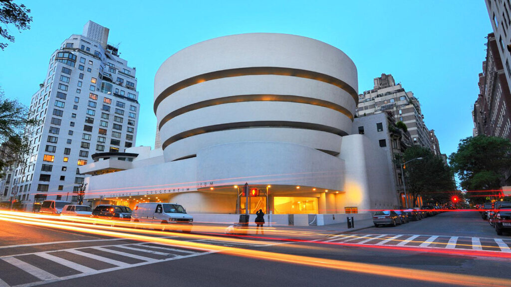 The Entrance to the Solomon R. Guggenheim Museum in New York City with Black Cars speeding By. Exterior view of the Solomon R. Guggenheim Museum on Fifth Avenue with black car service vehicles in front, highlighting luxury transportation to the museum. Main entrance of the Solomon R. Guggenheim Museum, showcasing its iconic curved façade and welcoming visitors to explore modern art.