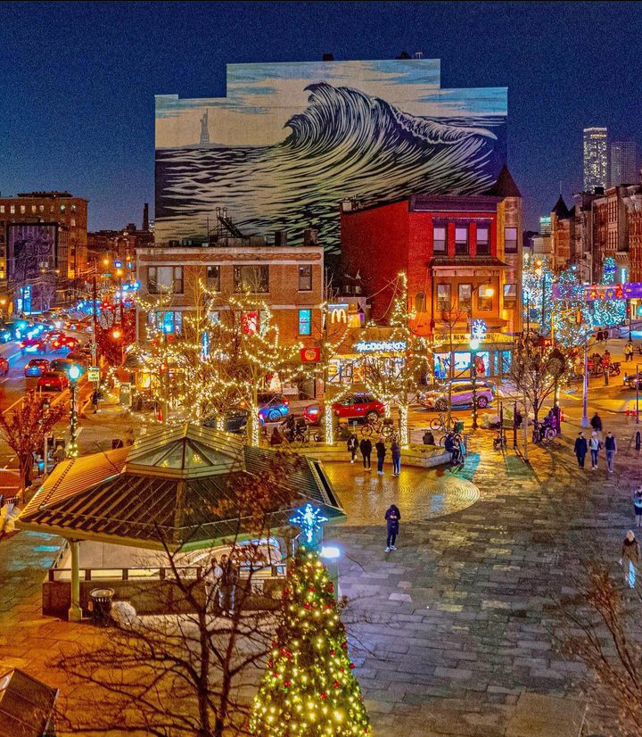 A vibrant holiday scene in Downtown Jersey City shows festive lights, busy streets, and a large mural overlooking the plaza, illustrating the lively atmosphere travelers experience when using private or long-distance transportation to or from New York City.