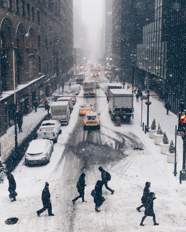 Snowfall blankets a busy New York City street as pedestrians cross a slushy intersection and yellow cabs, delivery trucks, and other vehicles navigate the slow, winter-weather traffic, highlighting the resilience of NYC transportation during harsh conditions.